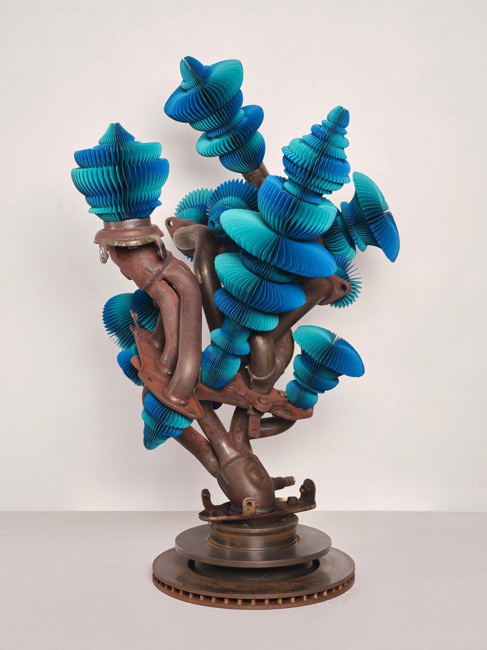 One of Miyazaki's car-muffler artworks: Dark metal car parts welded together so they look like the trunk and branches of a tree, with bright blue paper "flowers" attached to the ends