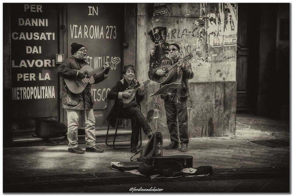 Image description: Three people play music on a corner in Naples, one guitar and tw mandolins.