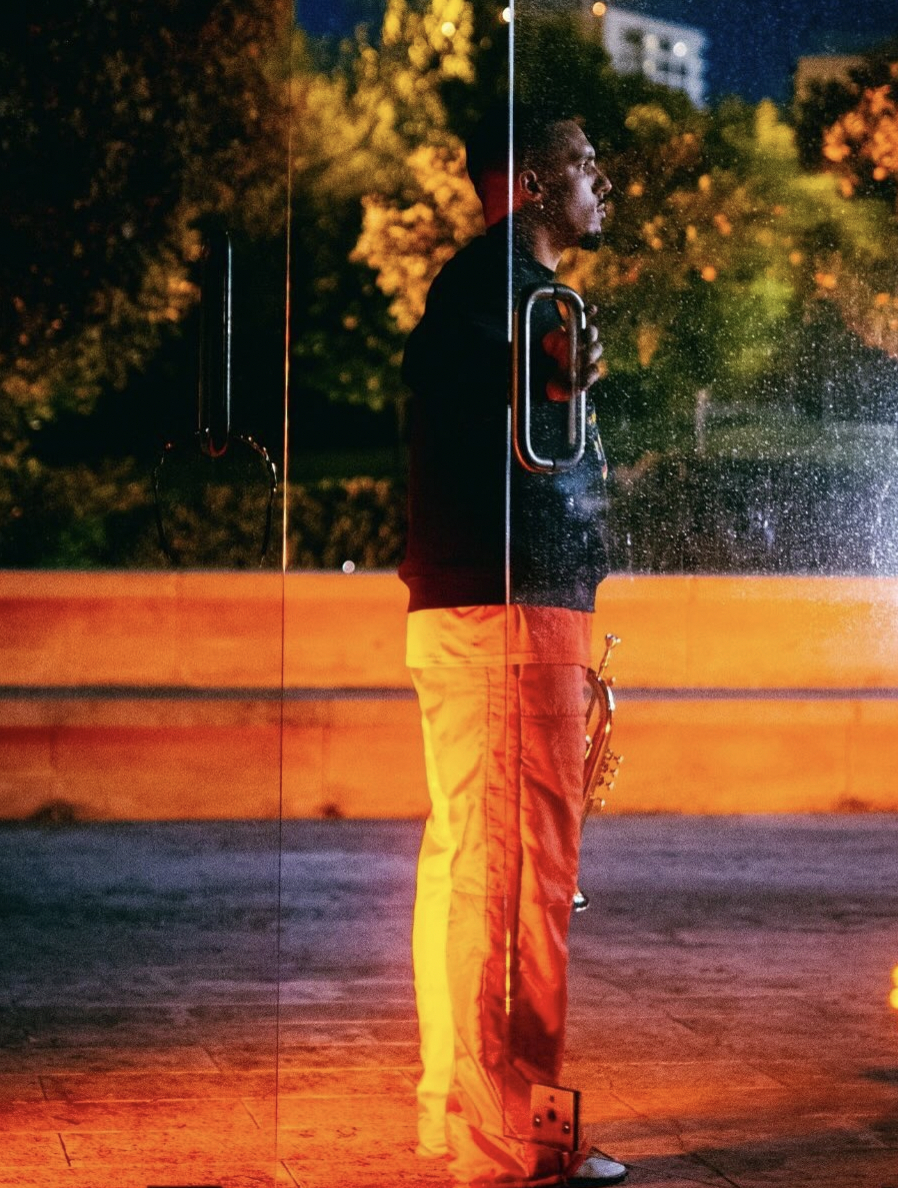 Tristany Mundu, bathed in orange light, wearing red trousers and black jumper, stands halfway out of a glass door in a glass wall leading to a patio holding a trumpet.