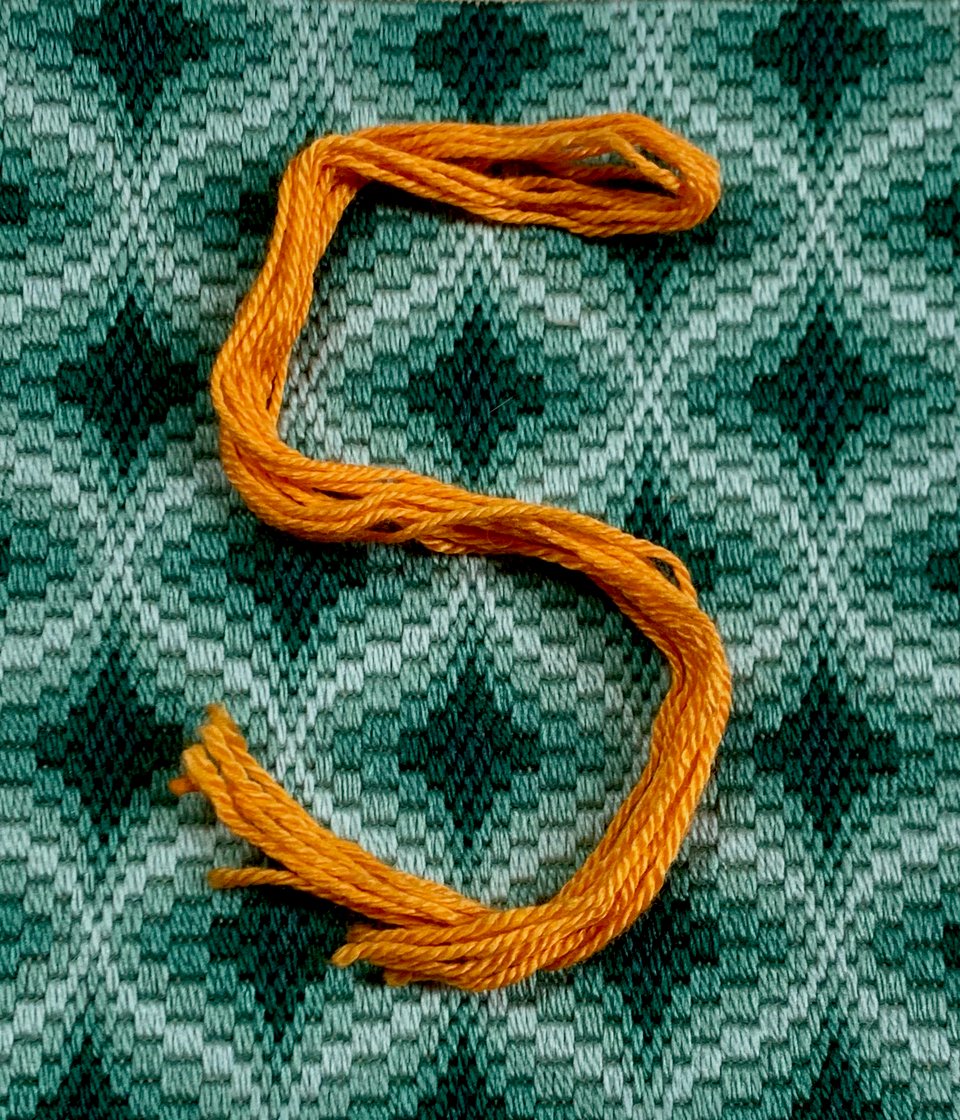 A bright orange hank of silk embroidery floss is arranged in the shape of a 5. In the background, there's a contrasting piece of bargello needlework in shades of olive green.