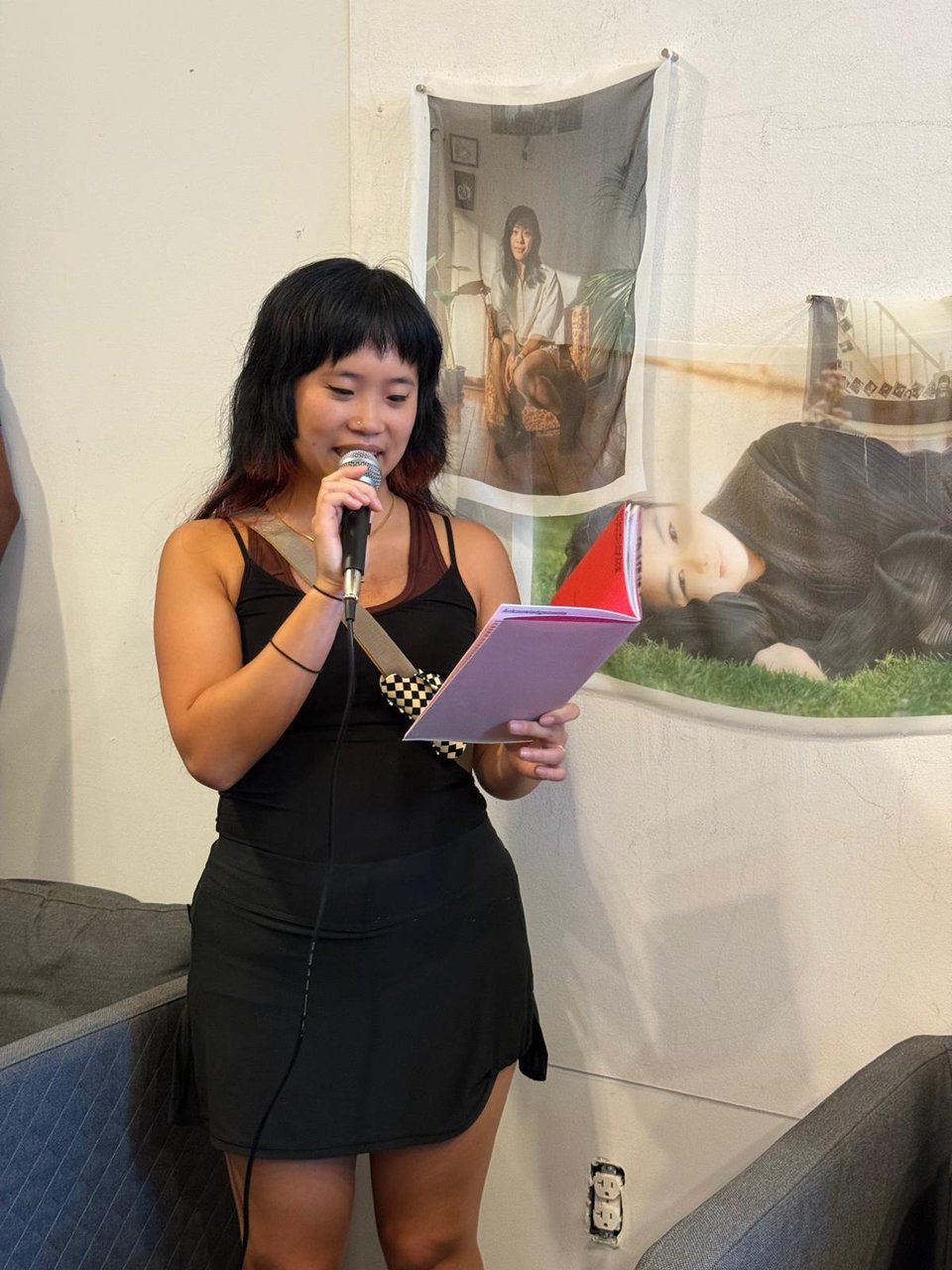 a chinese-malaysian person with long dark hair in a black dress reads from a zine, speaking into a microphone, behind them are portraits of asian women hanging on the wall printed on soft fabric