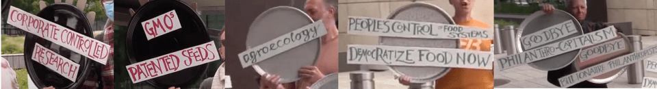 An array of protest signs, attached to foodservice trays, reading: Corporate Controlled Research; GMOs/Patented Seeds; agroecology; People's Control of Food Systems; Democratize Food Now; Goodbye Philanthrocapitalism; Goodbye Billionaire Philanthropists