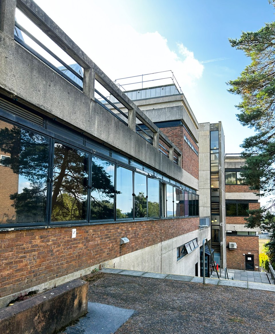 A building made up of several masses, going down a steep slope. The lower level of the nearest block is smooth concrete, with a band of redbricks and then a blackframed ribbon window surmounted by a line of black vents and topped by a concrete balustrade. Other blocks are more concrete.