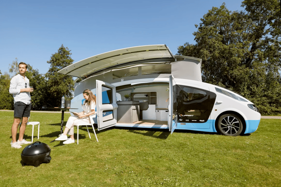 Photograph of a futuristic solar-powered camper van parked on a grassy field under a clear blue sky. The sleek white and blue vehicle has a bulbous roof and saloon-style doors open, revealing a modern interior with a compact kitchen and seating area. The roof is raised and lined with solar panels; extra slide-out panels extend sideways ou of the roof, creating shade for sittin beneath. A woman sits outside at a small table with a laptop, while a man stands nearby holding a drink. A portable black grill sits on the grass, and trees can be seen in the background, suggesting a relaxed and eco-friendly outdoor lifestyle.