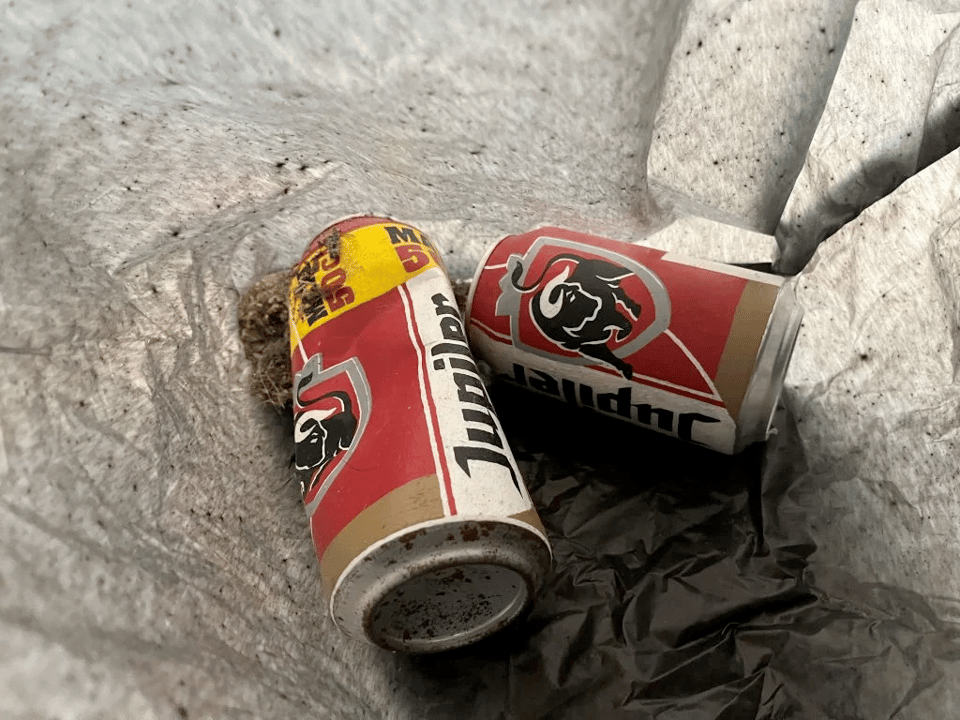 A photo of the two hand-painted cans. One is slightly larger than the other and is dented, while the other one is intact. They have been painted with a red and yellow design, and each sports a large logo of a black bull with white horns. The word "Jupiler" runs up the side of each can in a thick black font. It looks like a logo for this fictional beer