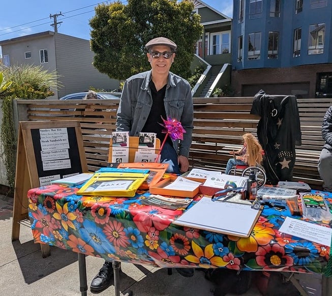 Vince in his sunglasses and flat cap, behind a table covered with colorful flowered vinyl, flyers, toolkits, and a wheelchair barbie