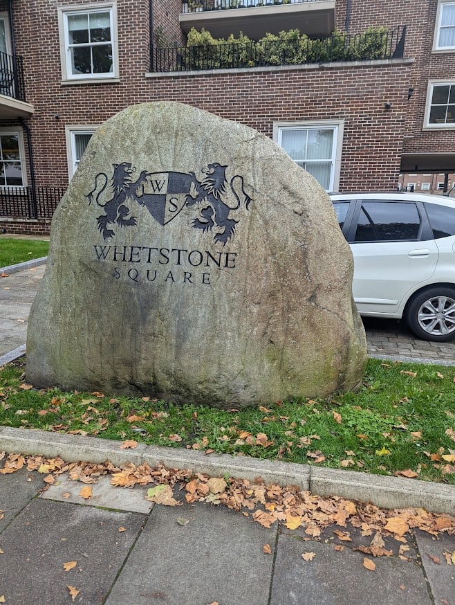 A big rock outside a housing block with a crest and the name 'Whetstone Square' engraved into it.