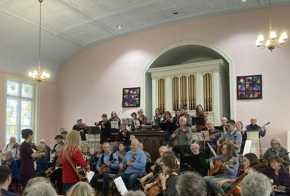 Musicians playing fiddles, guitars and mandolins play together at the front of a church.