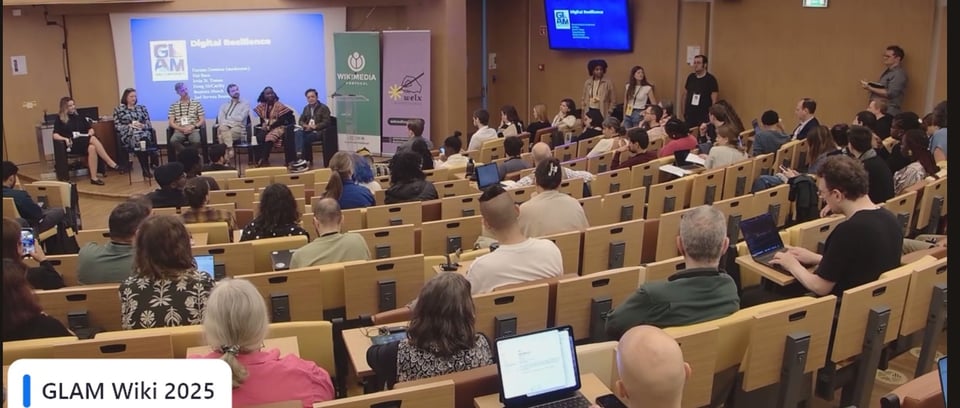 Six people sit on a stage on a panel with the auditorium filled with the audience.