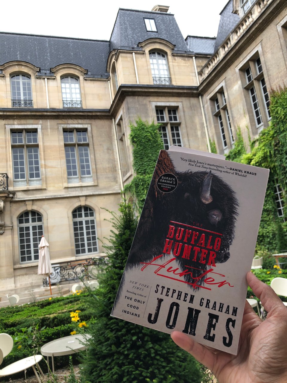My hand holding up a copy of The Buffalo Hunter Hunter in a (different) Parisian courtyard with elegant grey buildings, outdoor seating, low hedges, and creeprs on the walls.