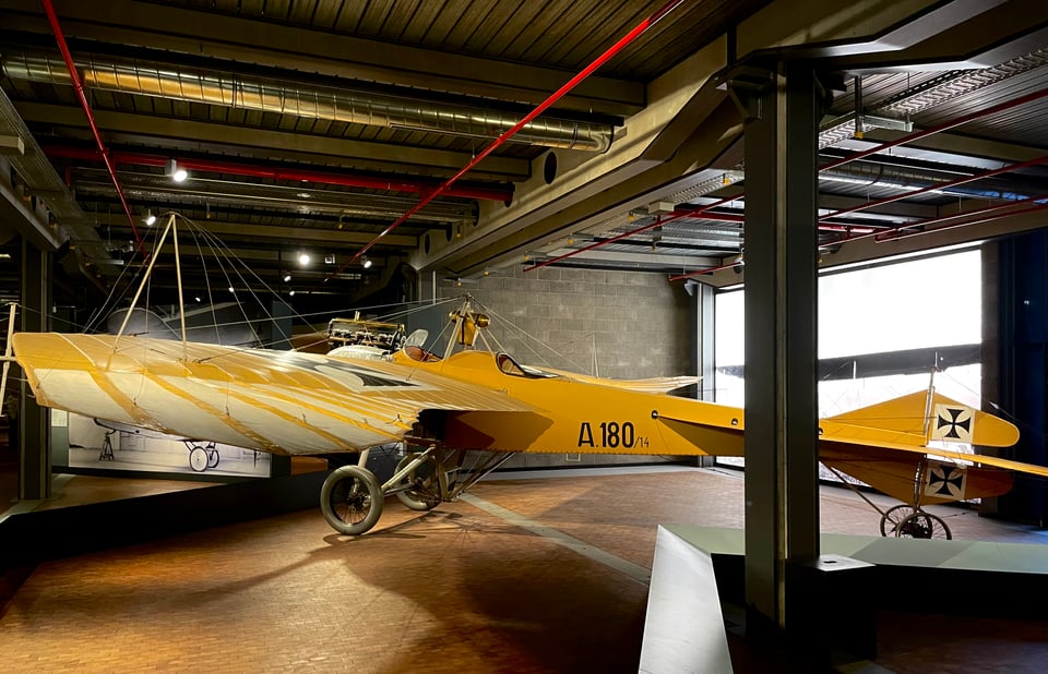 A Jeannin Stahltaube reconnaissance and training aircraft from 1914. Coloured lemon yellow, with white stripes on its wide wings. Wheels that look like from a fancy pram, and with a shiny steel nose (that is not visible in the current picture)