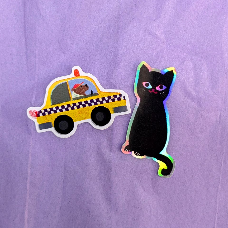 A photo of two stickers made from Kip's illustrations. One is a sticker of a bear driving a yellow taxi looking grumpy. The other is a holographic sticker of a black cat.
