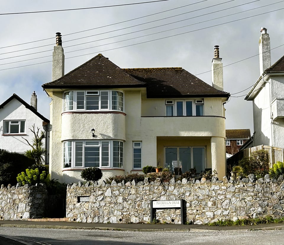 A detached suburban house. Although it is square set with a pitched rood, it has a large front bay with curved Critell style windows. At the right, a curved balcony forms a porch. It's so 1930s it could be in a early Vogue illustration.