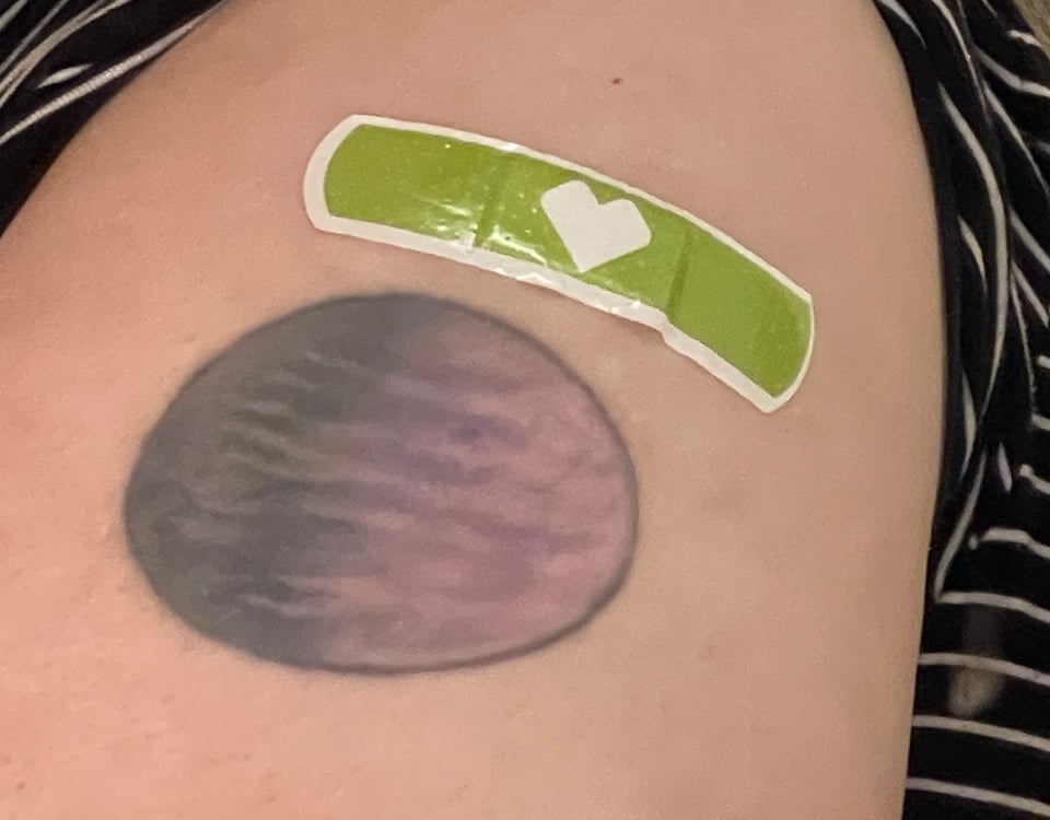 A spring-green bandaid with a white angular heart on it, taped onto a white person’s shoulder above a tattoo of a purple-and-black striped planet.