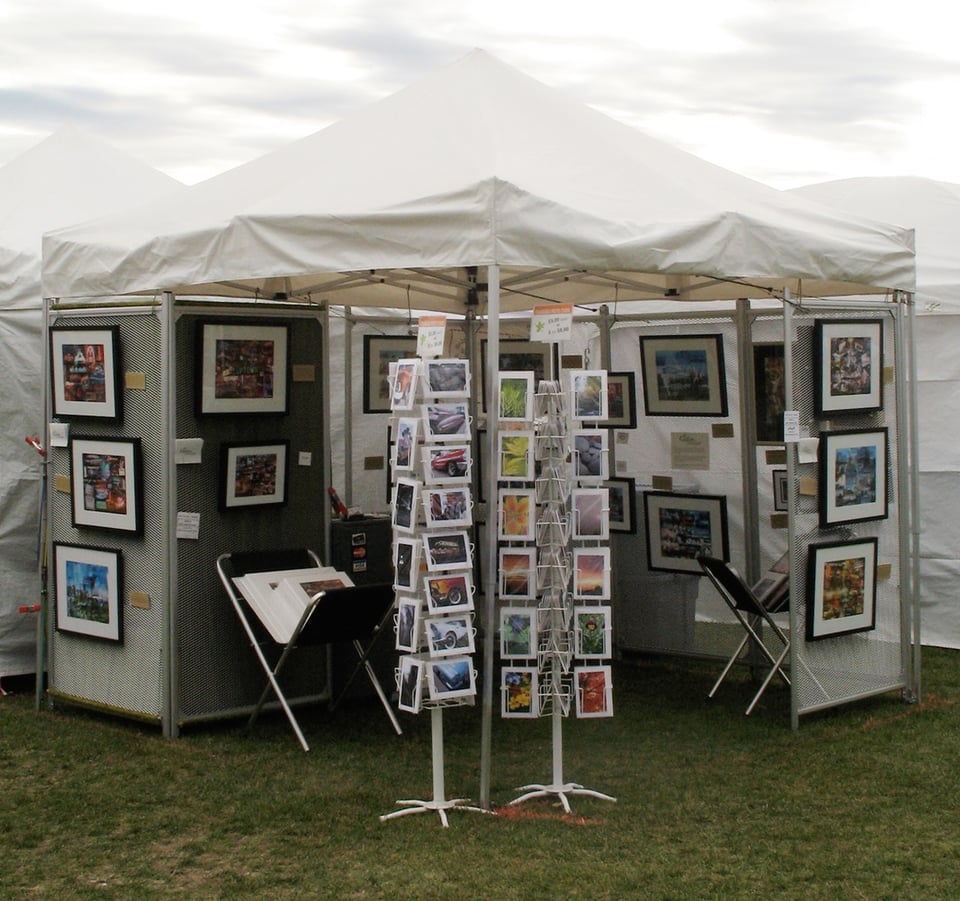 White tent at art fair with 2 card racks in front and folding displays with hanging framed art.