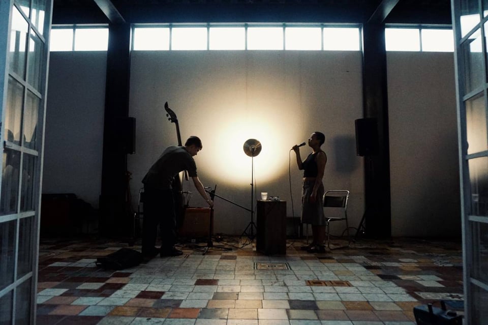 Theo and Caius are playing music from Crosspiece, singing and playing double bass. They face each other in a high ceiling room with multicolour tiled floor. Between them there's a bright lamp facing the wall.