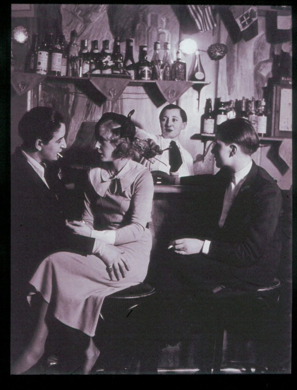 At the bar, a couple talks. The woman on the left is Lulu de Montparnasse, the owner of Le Monocle. She is in masculine dress and smoking a cigarette. She has her hand on the thigh of the woman she's speaking with.