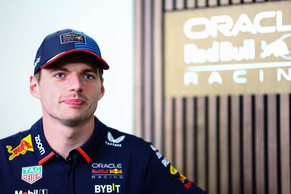 A comparatively happy Max Verstappen wearing his team kit, standing in presumbably a corporate team space with a monochromatic lit team logo behind him.
