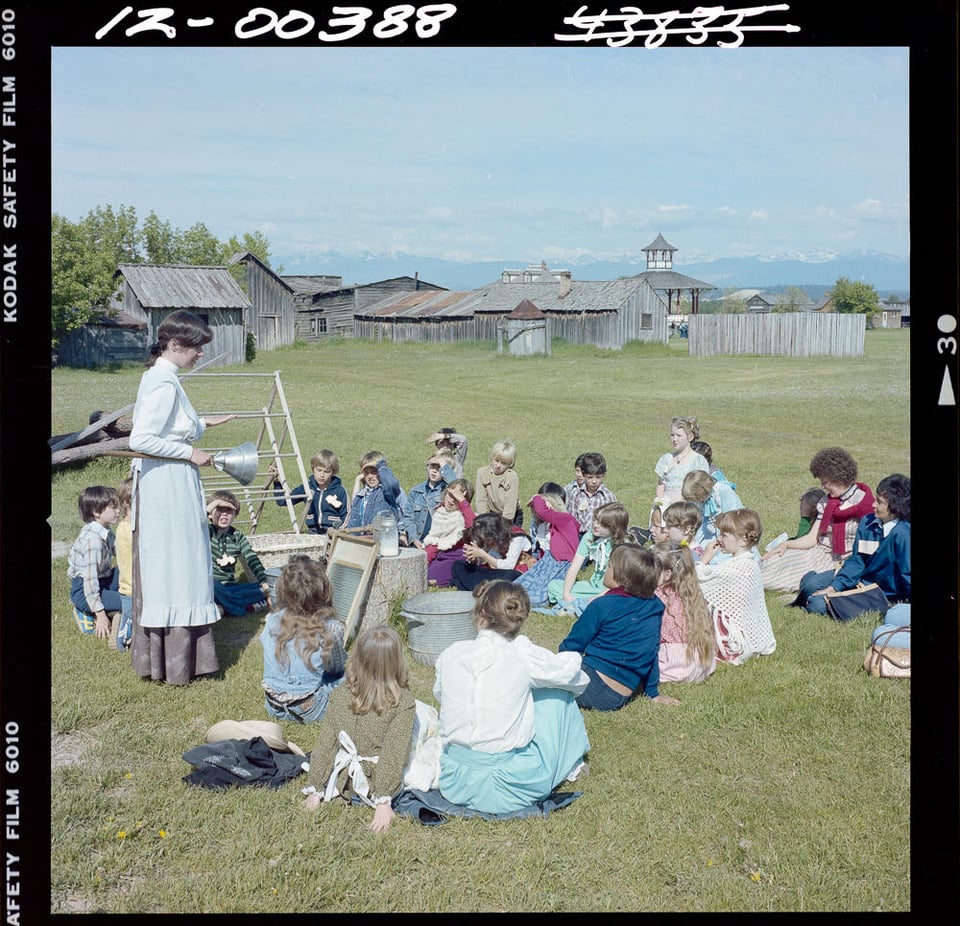 Colour photo. A group of children gather around a woman dressed in period costume holding objects that demonstrate old-timey laundry and washing.