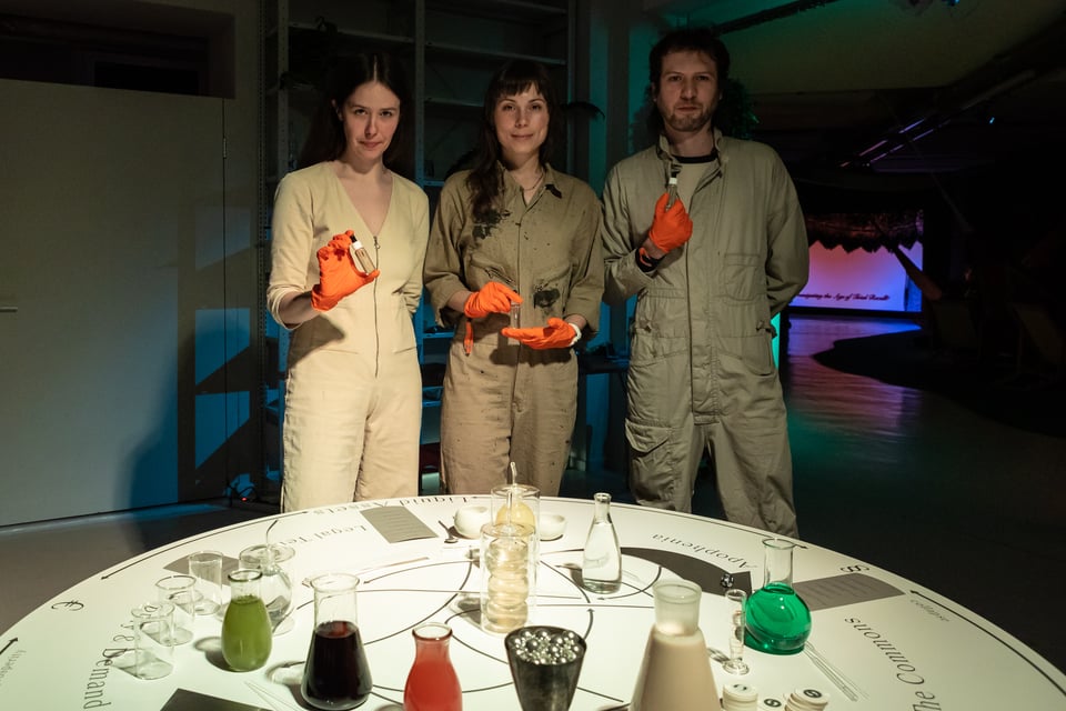 Gosia, Sarah and Arkadiy are standing (in that order from left to right) behind a table that holds a number of beakers and glass flasks, full of different coloured ingredients. They are all wearing beige jumpsuits and orange latex gloves, and holding small vials of syrup