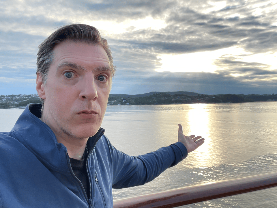A man with intense eyes points at a sunset on a fjord