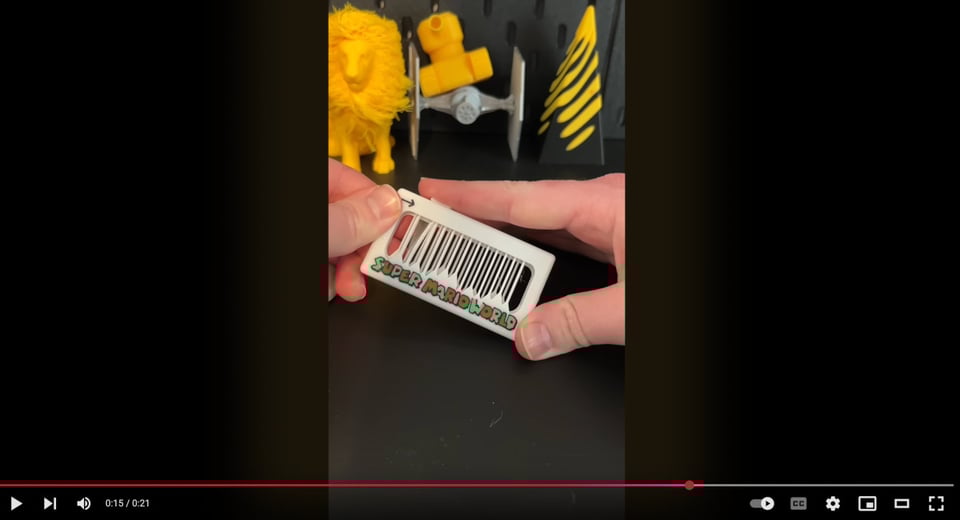 Screenshot of a YouTube video, showing two hands reaching out and holding one of the fidget toys, which is made of white plastic in a rectangular frame with an empty center, and perhaps it doesn't thin strips of plastic judging out through the center. It has the text "super Mario world" written on it in the classic super Mario font. In the background, there is a 3-D, printed lion, a 3-D printed tie fighter, and some sort of 3-D printed pyramid