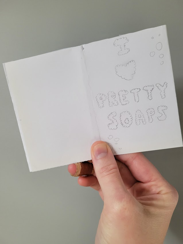 A white-skinned hand holding a mini-zine sketch with the title "I :heart: pretty soaps" in lettering that looks like bubbles.