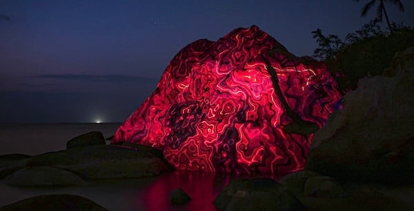 Una roca con curvas rojas y luminosas proyectadas en ella. Es de noche y contrasta con el cieño añil oscureciéndose.