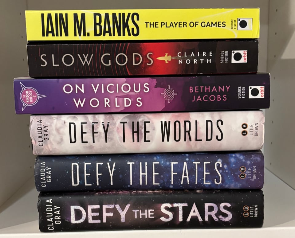 A stack of recently acquired books, spines out. Books: The Player of Games by Iain M. Banks, Slow Gods by Claire north, On Vicious Worlds by Bethany Jacobs, Defy the Worlds; Defy the Fates; and Defy the Stars by Claudia Gray.