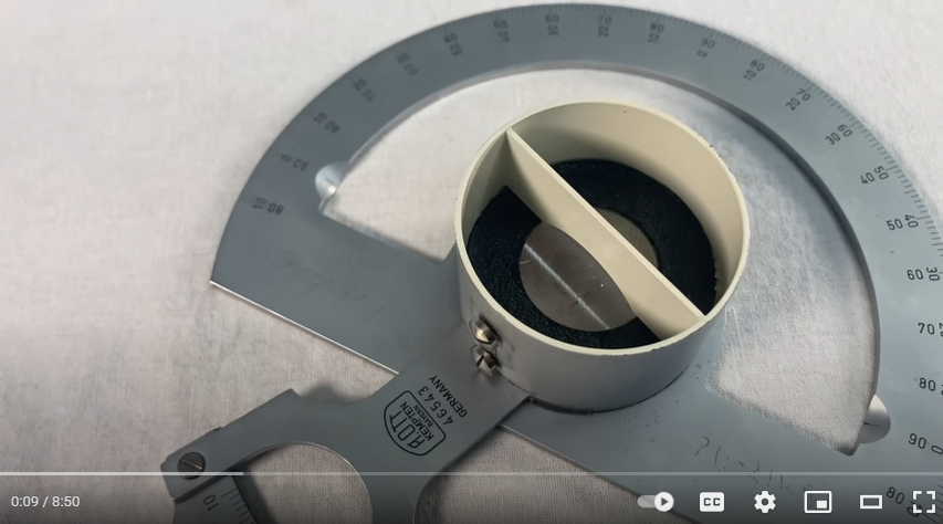A screenshot of a YouTube video focused on the “derivimeter”. It appears to be a metal protractor that has at its center a strange white cylinder bisected down the center, with a piece of glass near the bottom, adjacent to where the protractor lies on the paper. It is very cool looking.