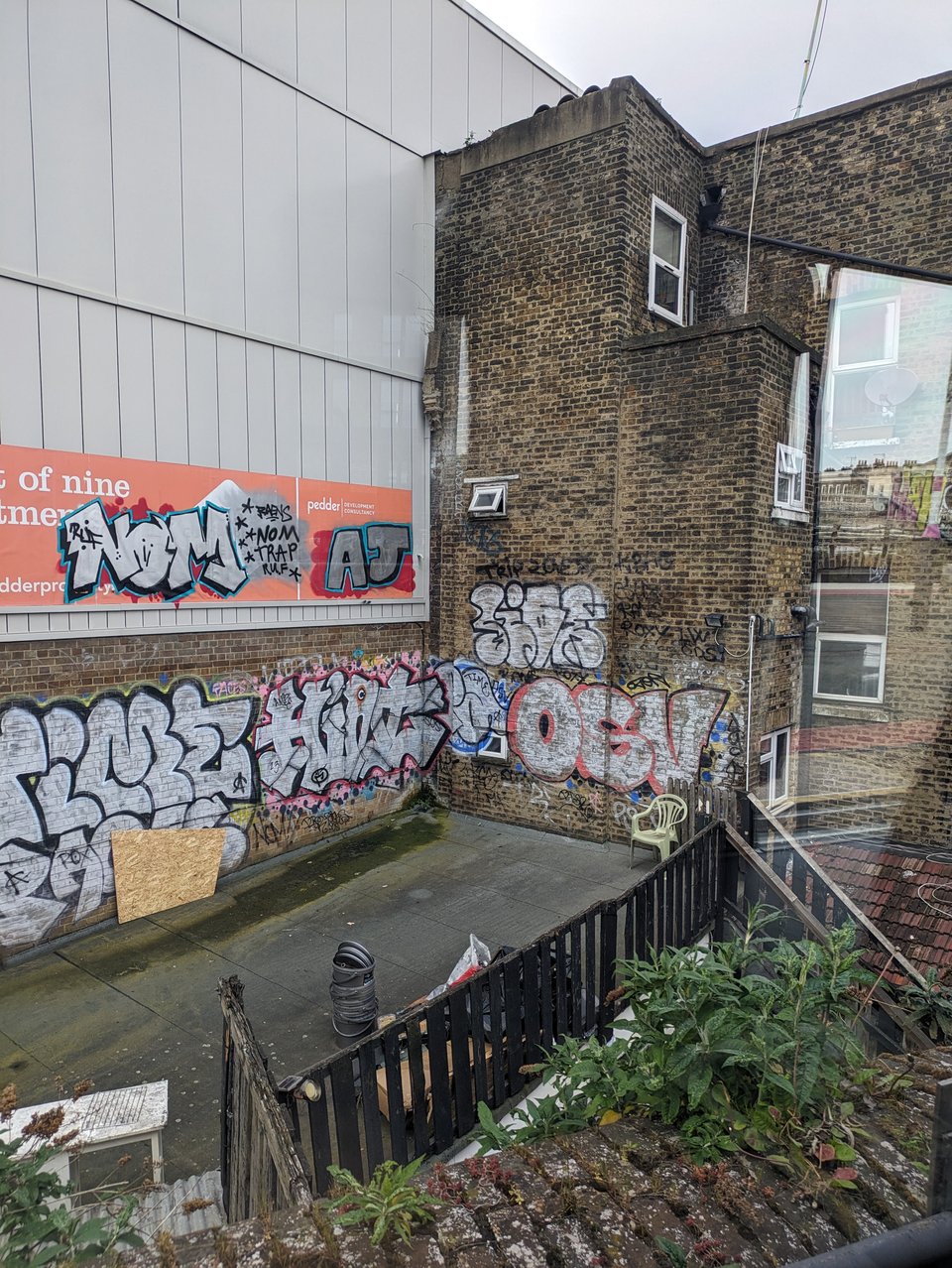 a view of a chaotic urban scene from a train, railings old brickwork, barbed wire and graffiti on a bland grey facade evoke the inspiring ugliness of London