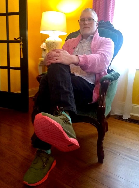 Andrew Neal sitting in a chair in 2020. He's chilling. Pink cardigan, green and pink shoes. Pretty slick haircut.