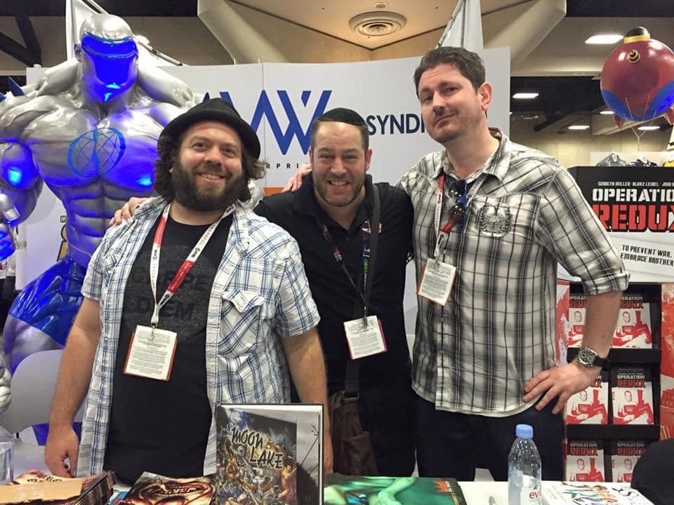 dan fogler, neil kleid and ben mccool smiling and standing a table with banners and a robot in the background