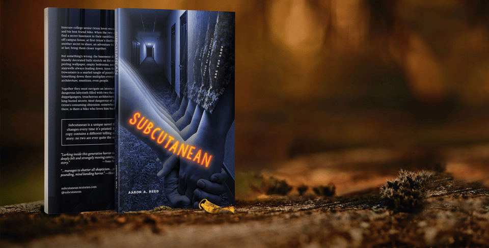 A picture of the novel Subcutanean, sitting on the dirt ground of what appears to be a cave or a basement. The book has a spooky blue image of a dark hallway with a closeup on the hands of two people who are holding hands. The title of the book floats in bright orange lettering