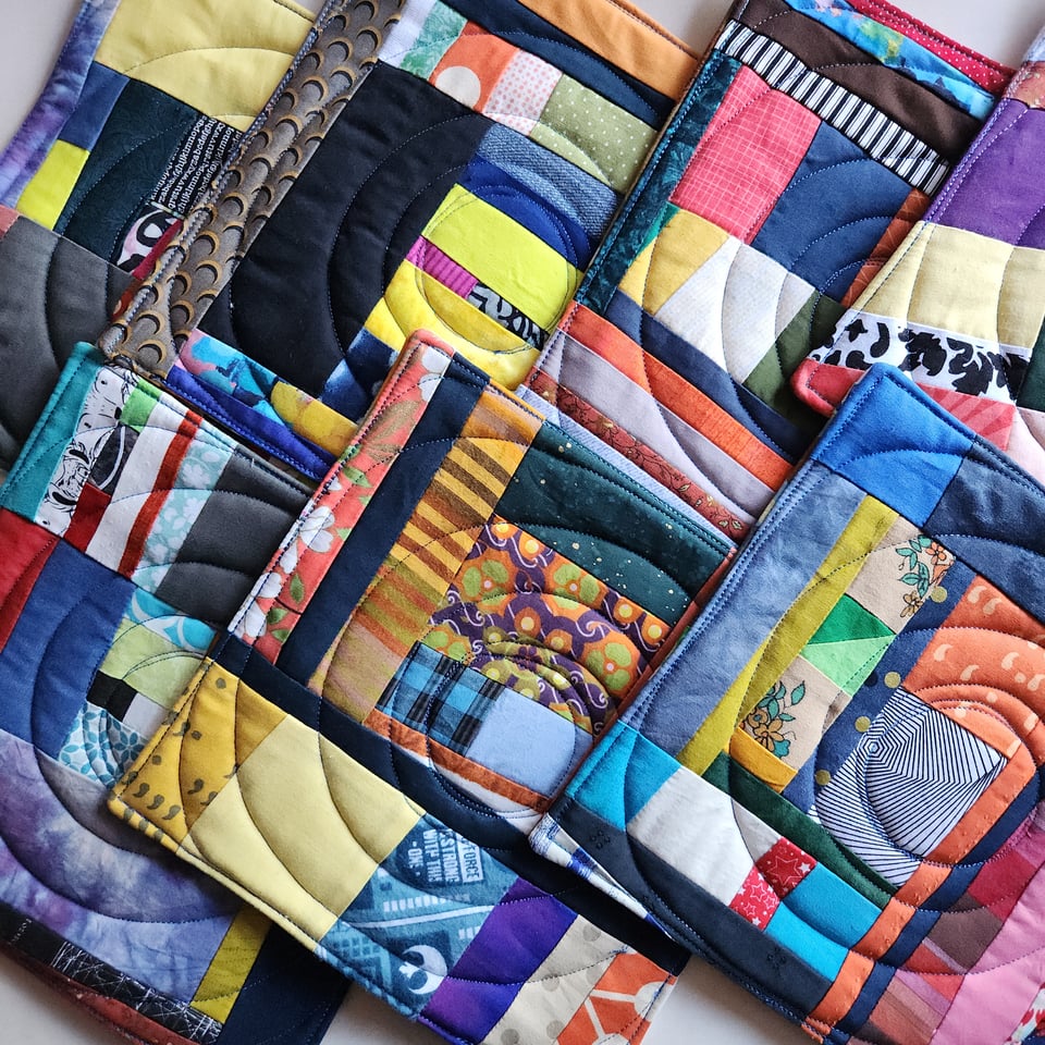 A pile of Sarah’s quilted potholders, using fabrics in all different colors, against a white fabric background.