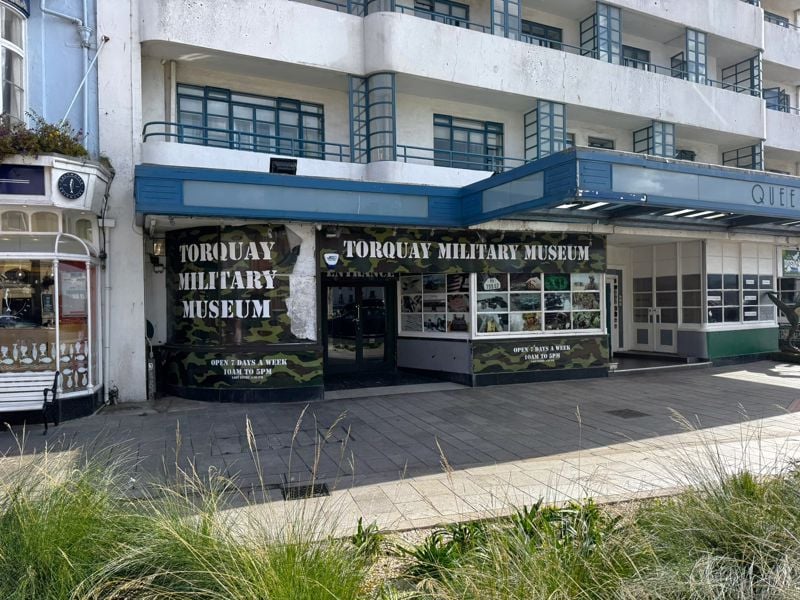 The left side of the frontage of the same Deco hotel. The projecting porch is in place, and the upper levels look the same, with the Crital frames now revealed as blue. The ground floor ribbon windows have had an entrance punched into them, and the windows mostly covered by a decal reading Torquay Military Museum over green camouflage.