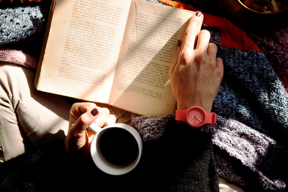Stock image of someone reading a book (face not shown) from an overhead angle - one hand is turning the page of a book while the other holds a cup of coffee or tea.