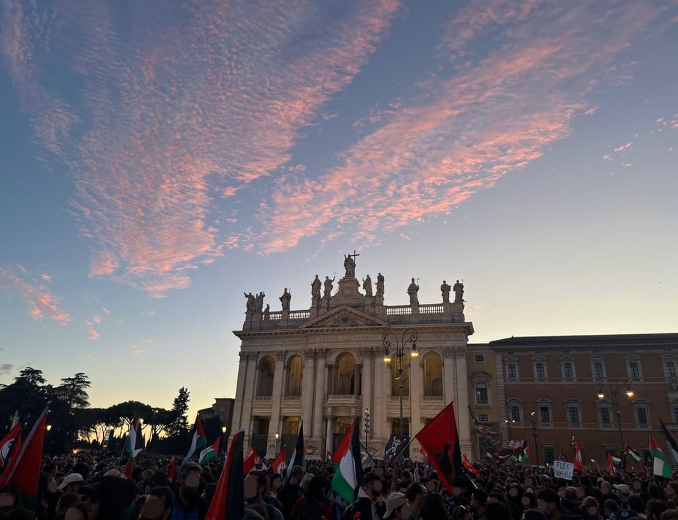 A large basilica is set against a dusk sky, with pink clouds. A crowd is visible at the bottom of the picture, with Palestinian and other red flags.
