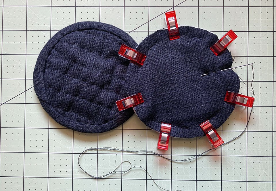 Two round, black linen coasters next to each other. One is completed with the hand quilting and binding done around the edges, and the other is in progress, with small red clips holding it together around the edges, and a needle is nestled into the fabrics, ready to begin the hand quilting.