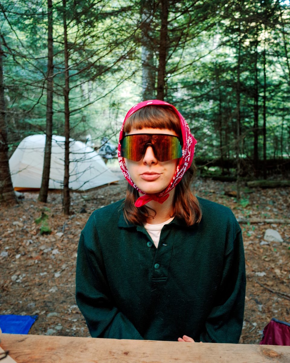 Sophie sits at a picnic table in the woods wearing silly sunglasses and a little scarf around their head. They are chewing.