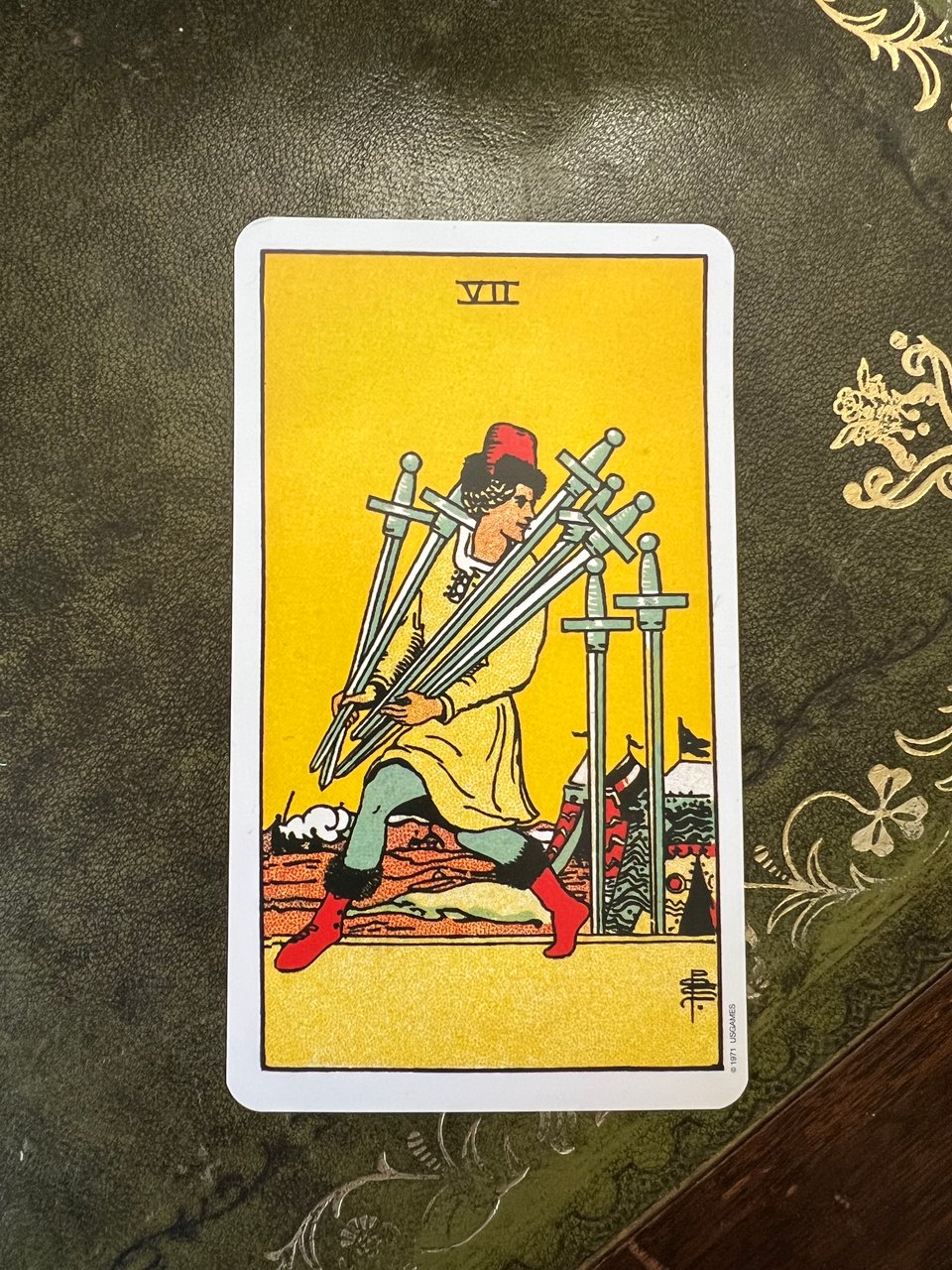 top down photo of the Seven of Swords tarot card on a vintage leather-topped desk. The card shows a man sneaking away from a military camp, carrying five swords and looking back longingly at the remaining two.
