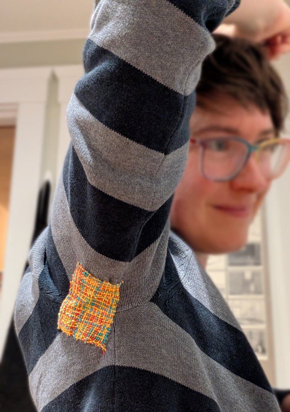 Maia lifts an arm to show off the darned patch on the armpit of her blue striped sweater. The patch is a stark contrast to the muted blue of the sweater, because it was made with a multicolored embroider floss that's primarily yellow, orange, and red. Maia's face can be seen out of focus in the background, and she looks pretty pleased with her darning job!