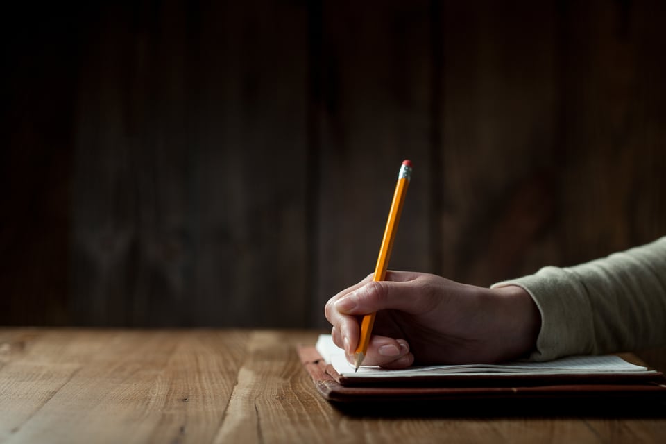 Stock image of a hand holding a pencil writing in a notebook on a wooden table.