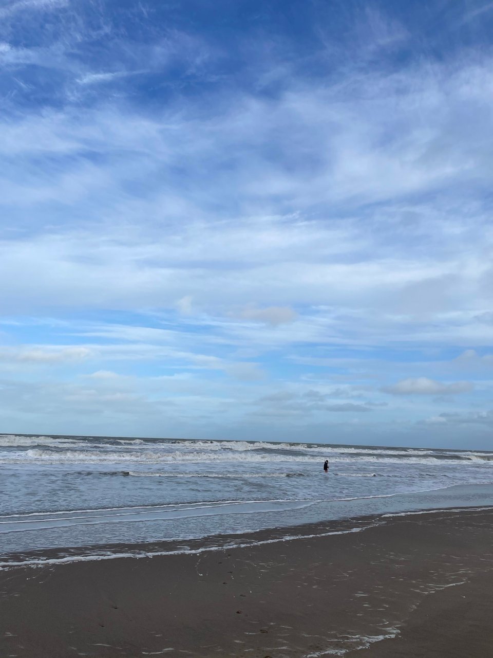 A lone man is waist high in the waves of ocean. He is tiny in the distance of the shot.