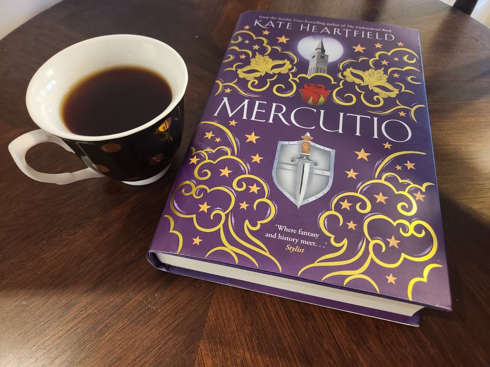 A harcover with a purple, gold and silver cover of Mercutio by Kate Heartfield, next to a cup of tea.