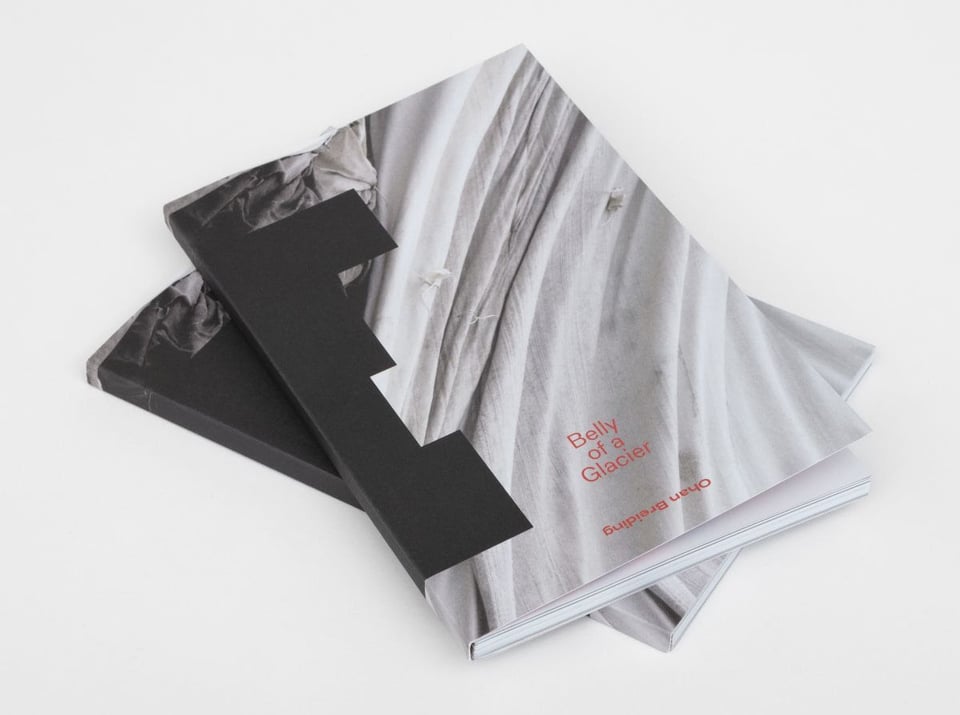 Two books stacked on top of each other with the title, Belly of a Glacier, and the name Ohan Breiding upside down, written in red. Cover image is a gray fabric and black rectangular shapes that envelope the book spine.