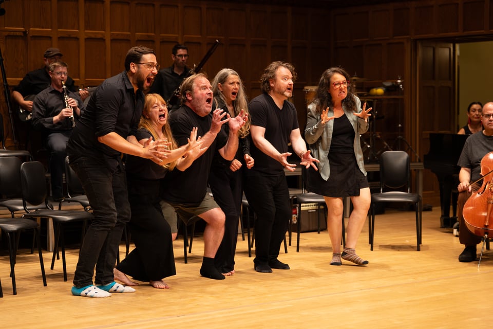 On stage, a group of actors screaming towards the audience, with some musicians visible in the background.