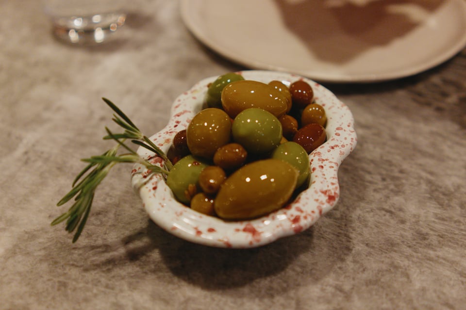 Various sizes and colors of delicious olives in a speckled serving dish with a sprig of rosemary.