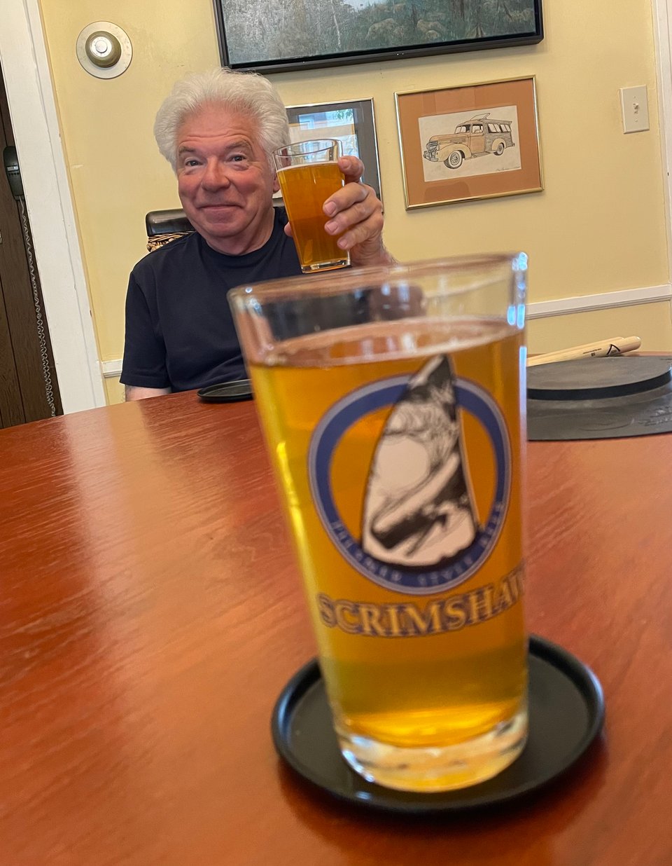 A beer in a pint glass labeled Scrimshaw is in the foreground. A man in the background smiles and lifts his glass in a cheers