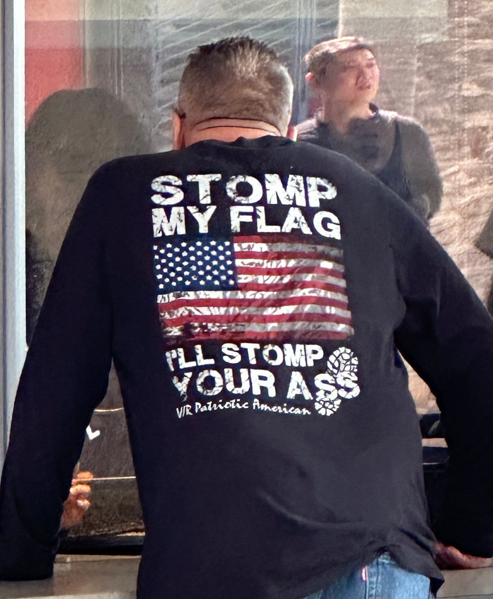 Photo of the back of a white man wearing a shirt that reads "STOMP MY FLAG I'LL STOMP YOUR ASS". He's at the ticket counter of the LA County Museum of Art.